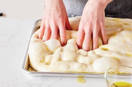 Billowy sheet of sourdough focaccia dough on a baking sheet being dimpled with a pair of hands.