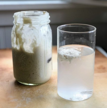 Beaker of water with a floating blob of sourdough starter, jar of starter in the background, illustrating the float test. 